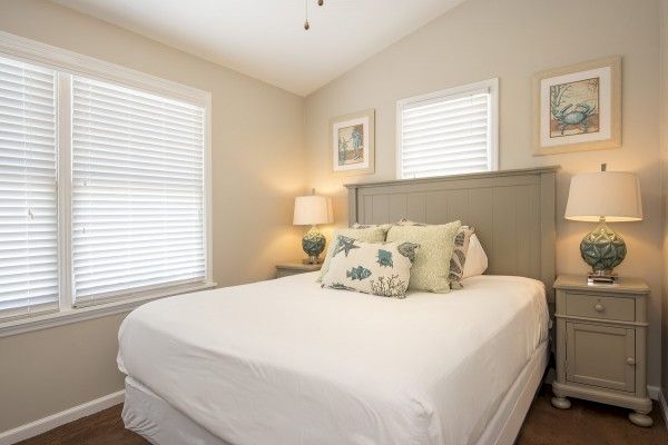 A bedroom with a bed, pillows, bedside tables, lamps, and framed artwork on the walls; two windows with blinds provide natural light.