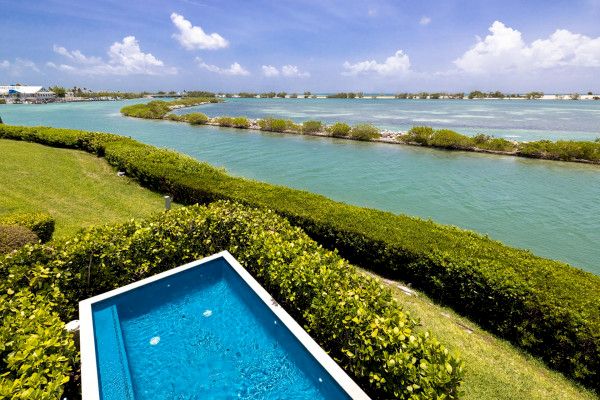 The image shows a bright, sunny landscape with a small pool in the foreground, surrounded by greenery, overlooking a tranquil body of water and islands.