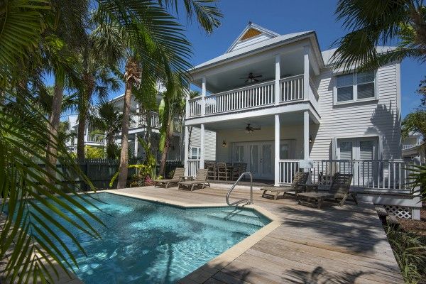 A two-story house with a pool in the backyard, surrounded by palm trees and outdoor furniture, including lounge chairs and dining tables.