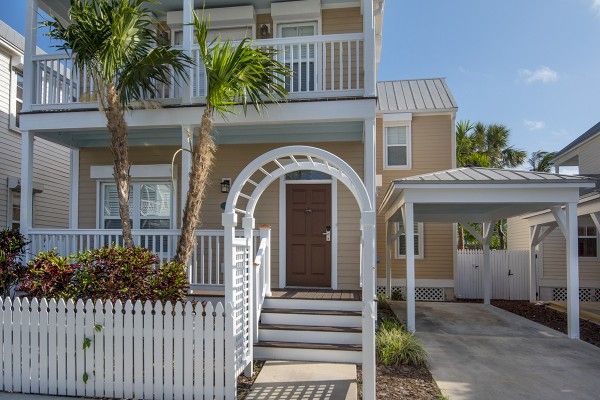 A two-story house with a white picket fence, front porch, and balcony; there's also a carport to the right and tropical plants around.