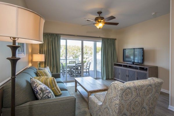 A cozy living room with a sofa, armchair, wooden coffee table, TV, and ceiling fan. Glass doors lead to a balcony with outdoor seating.