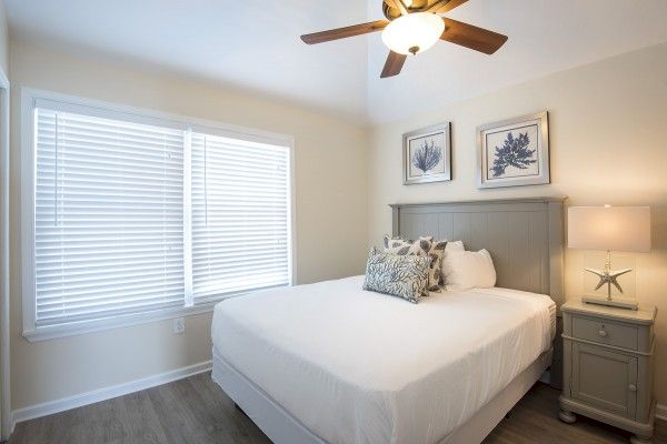 A bedroom with a bed, decorative pillows, two framed pictures above the headboard, a nightstand with a lamp, a ceiling fan, and a window with blinds.