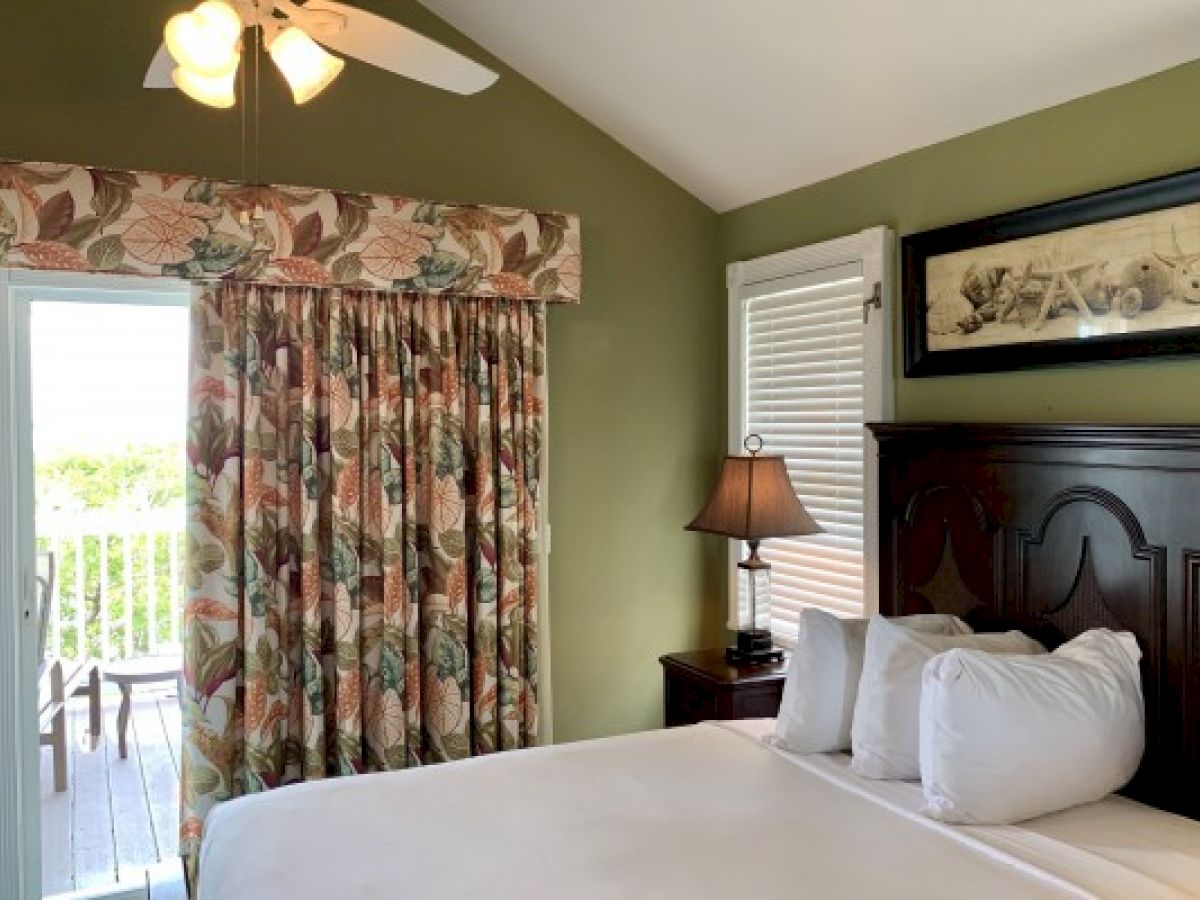 A cozy bedroom with green walls, floral curtains, a ceiling fan, a lamp, and a bed near a door leading to a balcony.