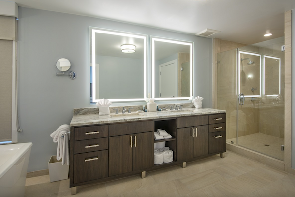 A modern bathroom with a double sink vanity, illuminated mirrors, a glass-enclosed shower, and a bathtub, all featuring sleek and elegant design elements.