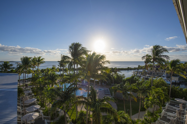 This image shows a scenic view of a tropical resort with lush palm trees, a swimming pool, and the sun setting over the ocean in the background.