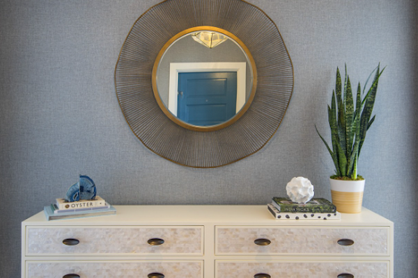 A stylish console table with four drawers, topped with decorative items, sits beneath a circular mirror and a potted plant on a gray wall.