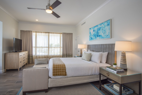 A modern bedroom with a bed, nightstands, lamps, a wall art piece, a TV on a dresser, and a large window with curtains ending the sentence.