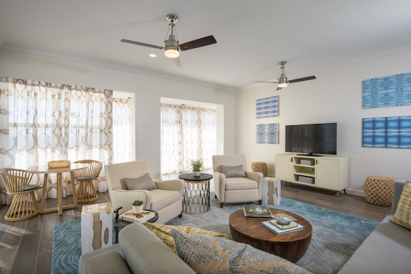 A modern living room with neutral colors, featuring sofas, armchairs, a round coffee table, TV, and abstract wall art. Two ceiling fans are installed.