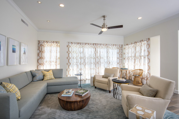 A modern living room with a gray sectional sofa, beige armchairs, a wooden coffee table, patterned curtains, and wall art, well-lit by natural light.