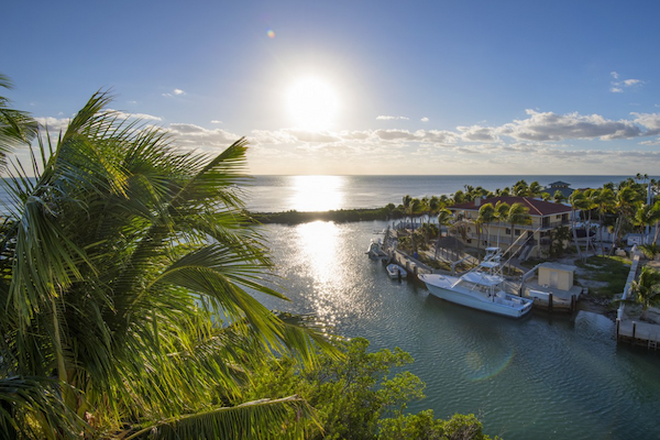 A coastal scene at sunset with palm trees, a waterway leading to the ocean, boats docked near houses, and a clear blue sky.