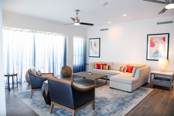 A modern living room with a sectional sofa, armchairs, a coffee table, framed artwork, and a carpeted floor, lit by natural light through large windows.