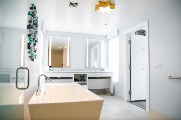 A modern bathroom with a large white bathtub, dual sinks, mirrors, and decorative wall art. The room is bright and minimalistic.