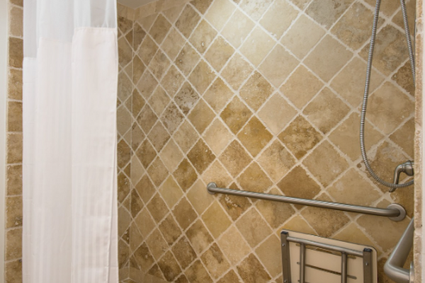 A walk-in shower with beige tiles, a white curtain, handheld showerhead, metal grab bar, and folding shower seat.