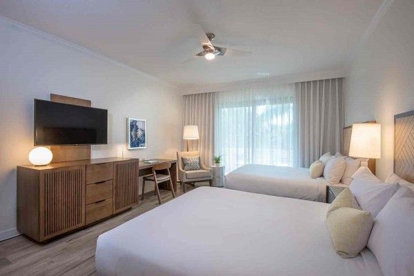 This image shows a neatly arranged hotel room with two beds, a TV on a wooden cabinet, a desk with a lamp, and a seating area by the window.