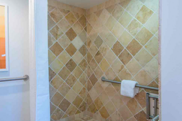 The image shows a walk-in shower with beige tiled walls and floor, a curtain rod with a white curtain, a grab bar with a towel, and recessed lighting.