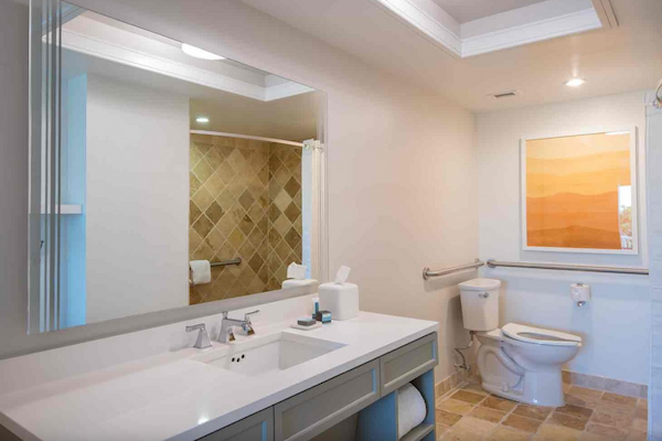 A modern bathroom with a large mirror, sink, toiletries, toilet, and a walk-in shower with a tiled wall and a piece of artwork on the wall.