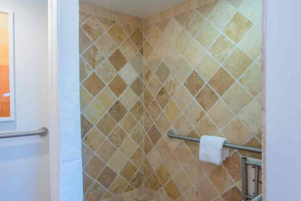 The image shows a tiled walk-in shower with a white curtain, safety handrails, and a towel area. The tiles are in a beige and brown color scheme.