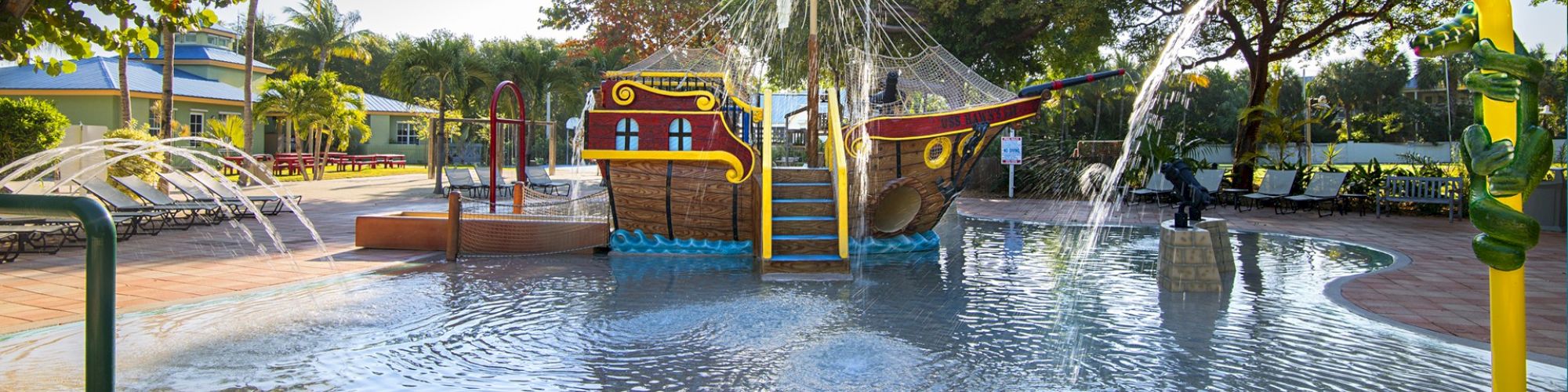 A colorful water playground featuring a pirate ship, water fountains, and splash zones, set in a park with trees and surrounding benches.