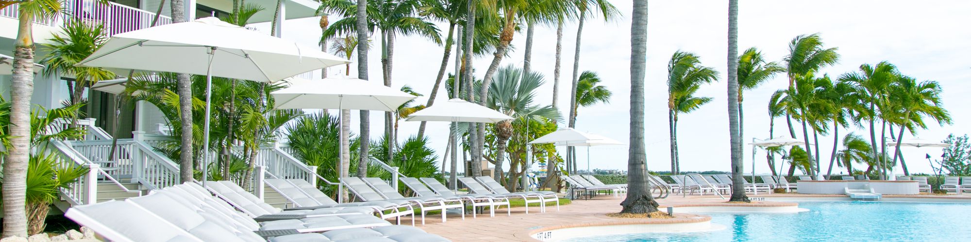 A poolside area with white lounge chairs, umbrellas, and palm trees, near a building under a partly cloudy sky.