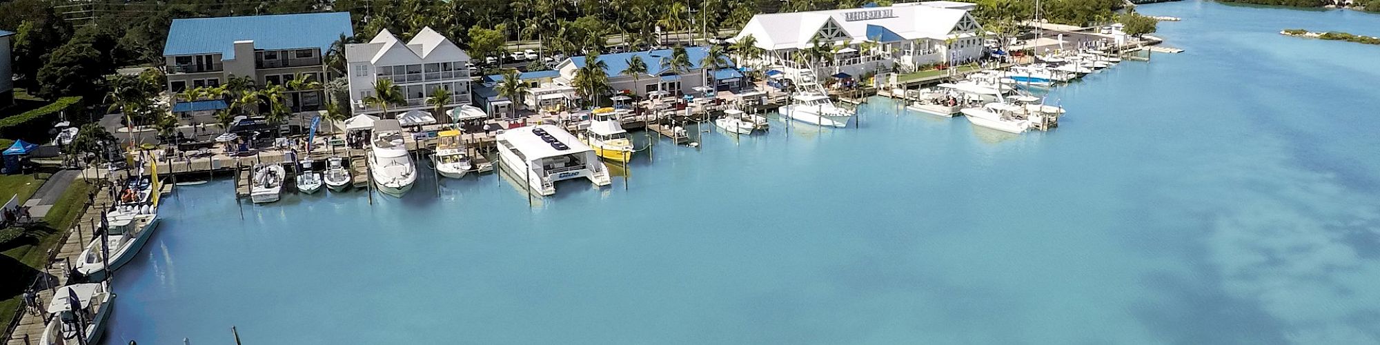 A picturesque marina with clear blue water, docked boats, and waterfront buildings, possibly a resort or residences, surrounded by lush greenery.