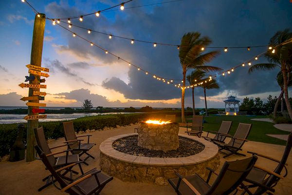 A cozy fire pit surrounded by chairs, string lights, and a palm tree, with a scenic outdoor backdrop at dusk, near water and a signpost.