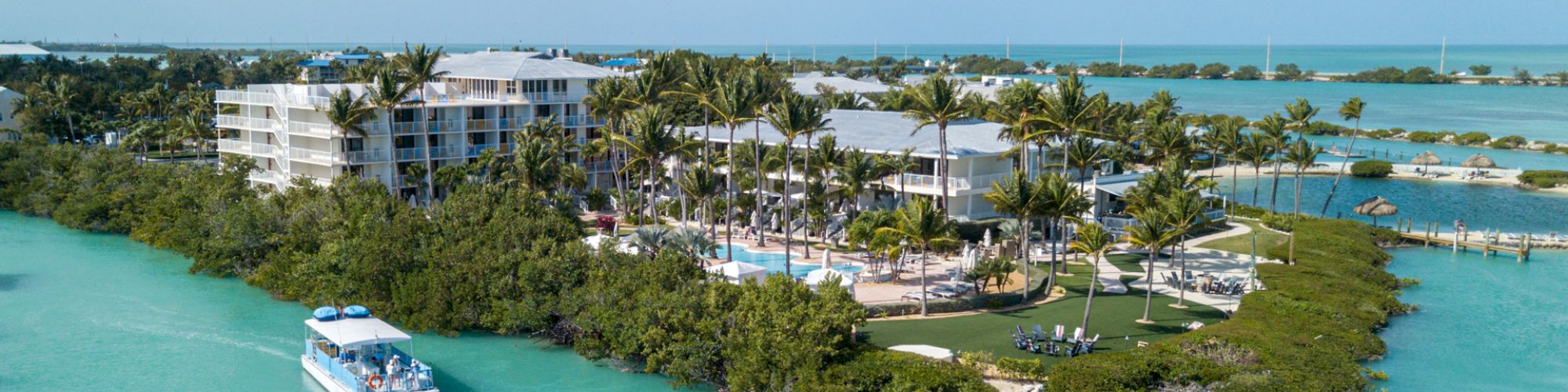 A tropical resort with palm trees, buildings, and a boat in turquoise water, surrounded by lush greenery and clear skies in the background.