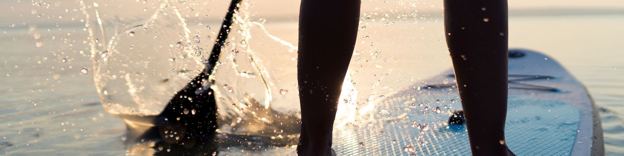 A person stands on a paddleboard, with water splashing from the paddle under a clear sky.