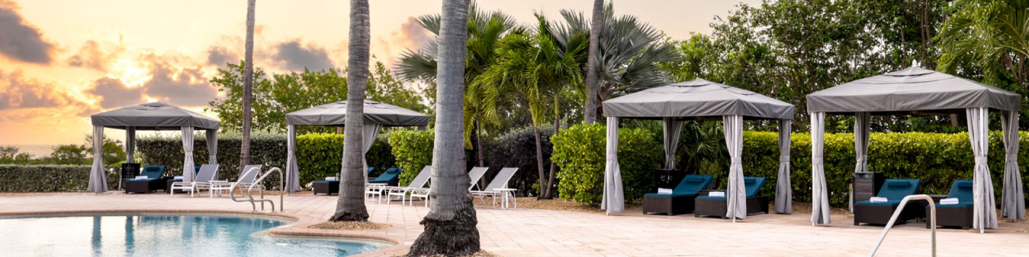 The image shows a serene pool area with lounge chairs, cabanas, and palm trees set against a sunset sky, surrounded by lush greenery.