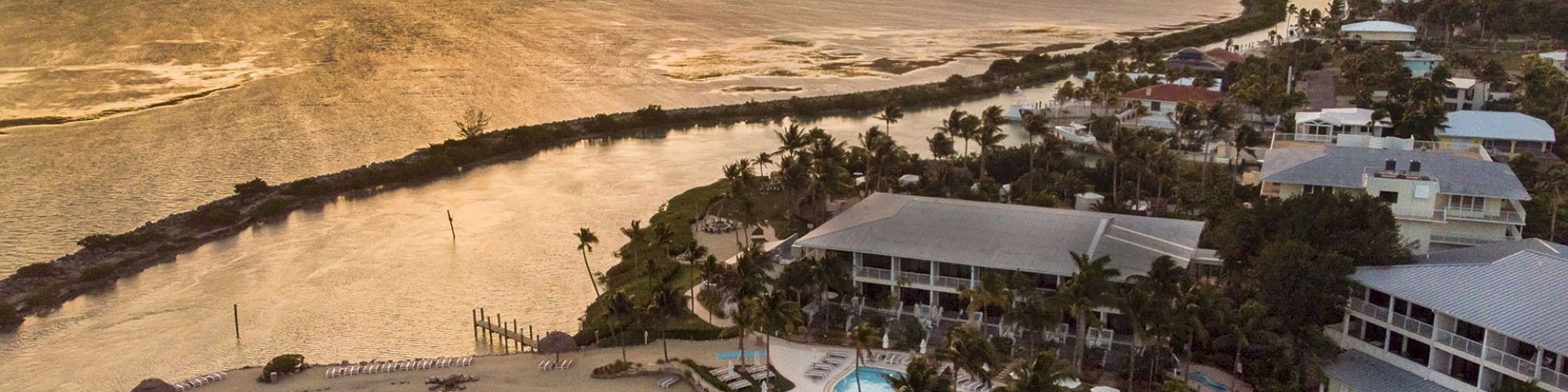 A coastal resort area at sunset, featuring a hotel with pools, palm trees, a sandy beach, and water bodies leading to the ocean.