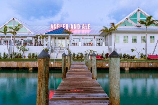 A wooden dock leads to a building with a neon sign reading "ANGLER AND ALE," flanked by palm trees and surrounded by water.