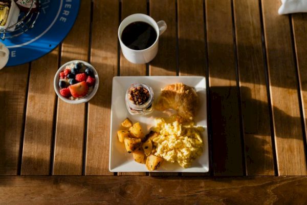 A breakfast spread on a wooden table with scrambled eggs, potatoes, croissant, yogurt with granola, a cup of black coffee, and a fruit bowl.