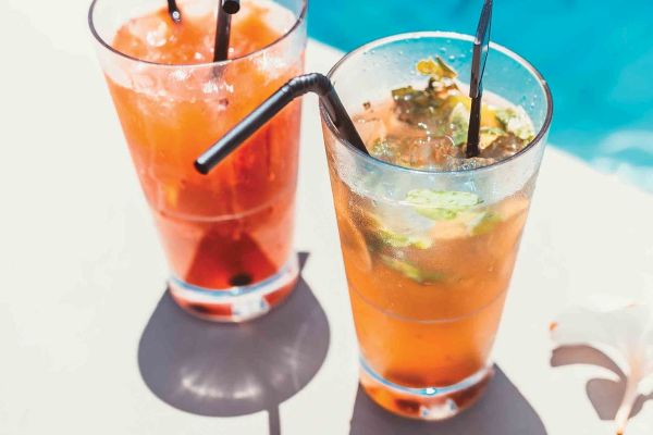 Two glasses of beverages with straws on a poolside table, accompanied by a white flower, set against the clear blue water.