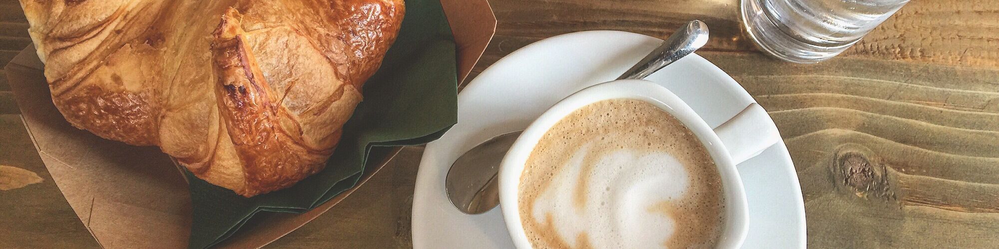 A croissant on a tray, a cup of coffee with a saucer and spoon, and a glass of water on a wooden table.