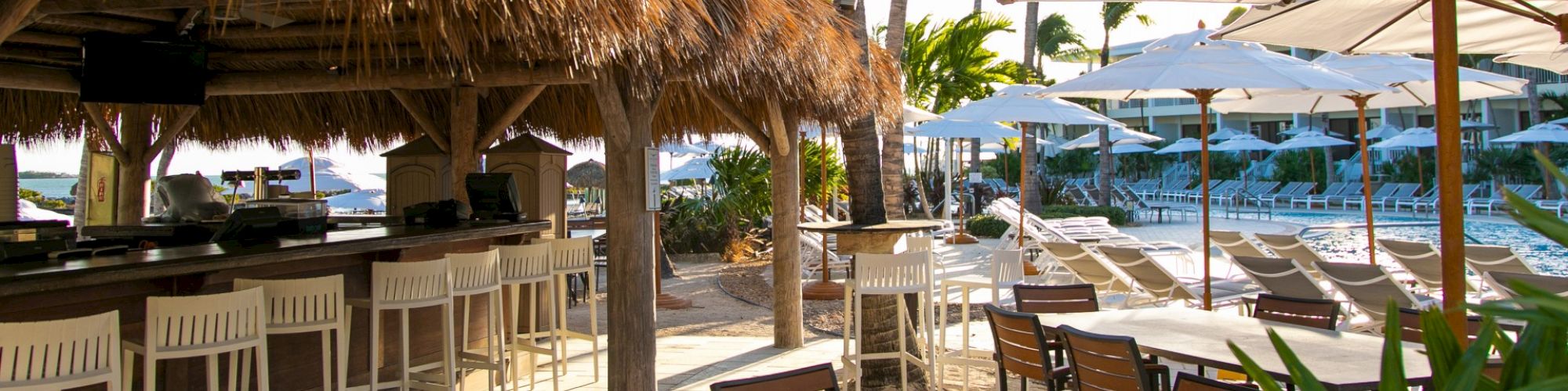 The image shows an outdoor resort area with a tiki bar, white bar stools, dining tables, chairs, and loungers beneath umbrellas next to a pool.