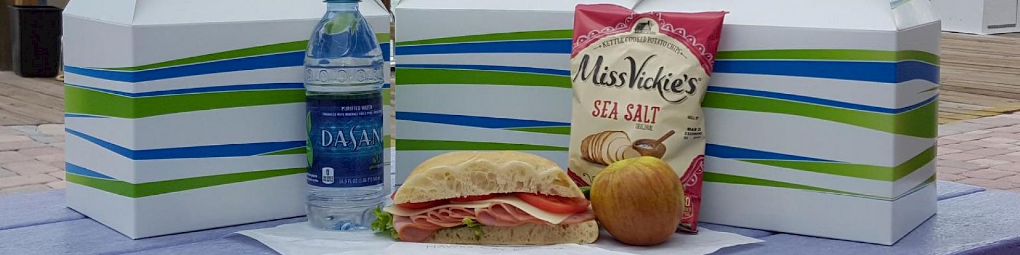 Three box lunches with a bottle of water, Miss Vickie's Sea Salt chips, a sandwich, and an apple on a table.