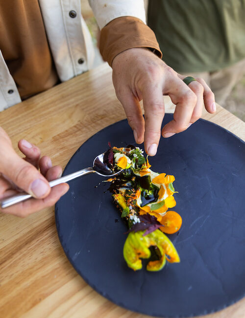 A person is arranging colorful, artistic food on a dark plate with a fork and hand, over a wooden table surface.