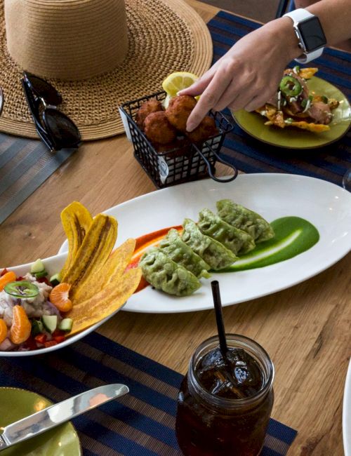 A table with a variety of dishes, including guacamole, salad, fried snacks, drinks, and a person reaching for food, is depicted in the image.