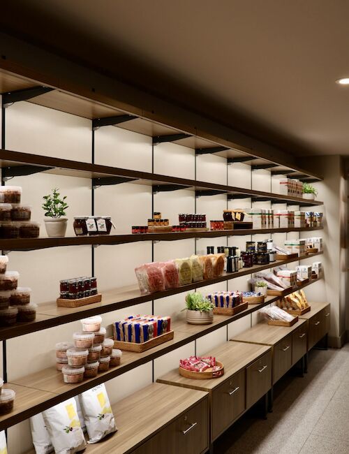 The image shows a neatly organized display of snacks, drinks, and other items on wooden shelves in a modern store or pantry setting.