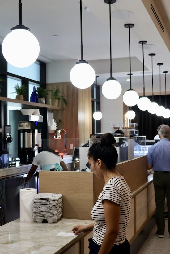 A modern cafe with pendant lights, a marble counter, and people in line near a wooden wall and black curtain.