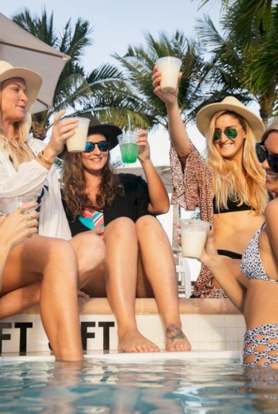 A group of women in swimsuits and hats are enjoying drinks by a poolside, surrounded by palm trees and an umbrella.