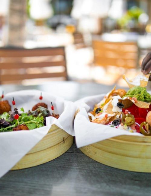 Two people are reaching for food in round wooden trays outdoors; the food includes a variety of appetizers and salads.