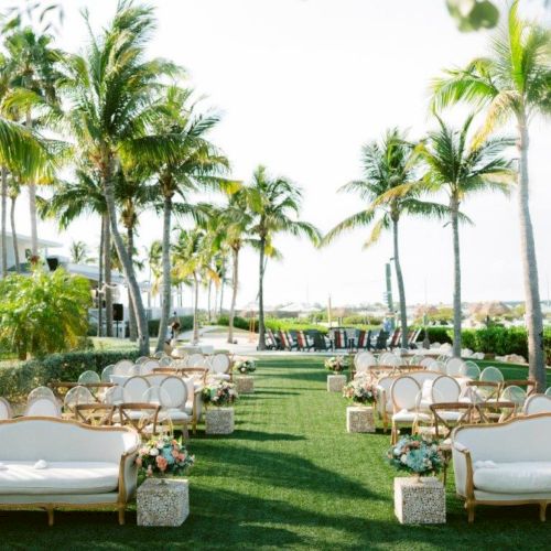 A beautifully decorated outdoor wedding setup featuring elegant white chairs and sofas, with a backdrop of palm trees and a waterfront view.