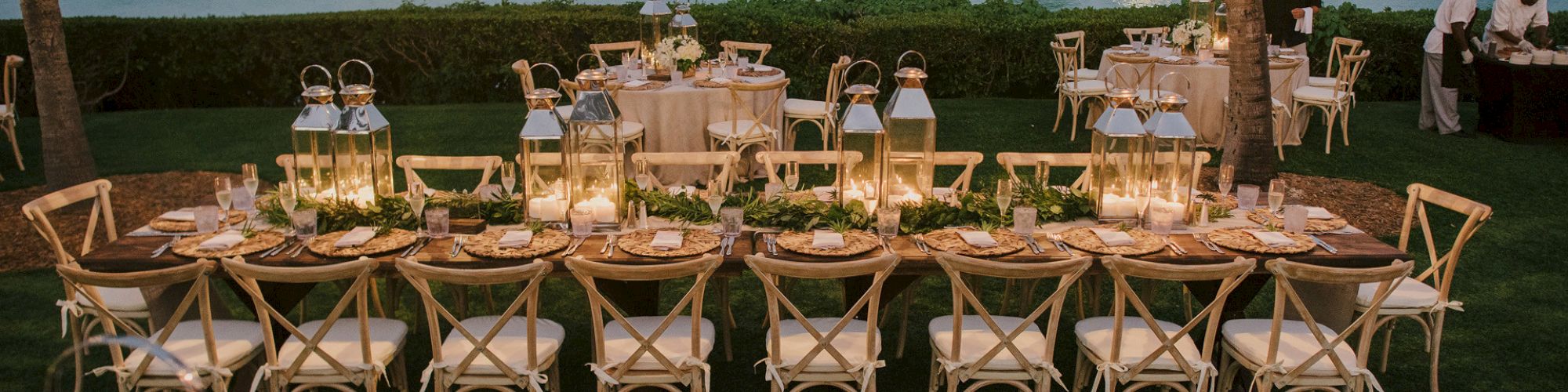 An elegant outdoor dining setup by a waterfront, featuring long tables with rustic decor, lanterns, and ambient lighting, ready for a special occasion.