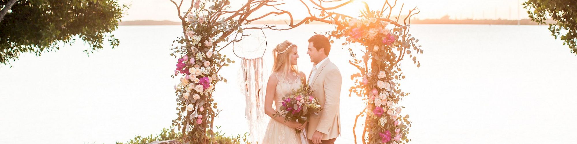 A couple stands under a decorated arch on a beach, facing each other, surrounded by empty chairs. The setting looks like a wedding ceremony.