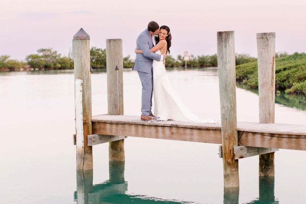 A couple stands at the end of a wooden dock over calm water, sharing a kiss with their reflections visible below, against a serene sky backdrop.