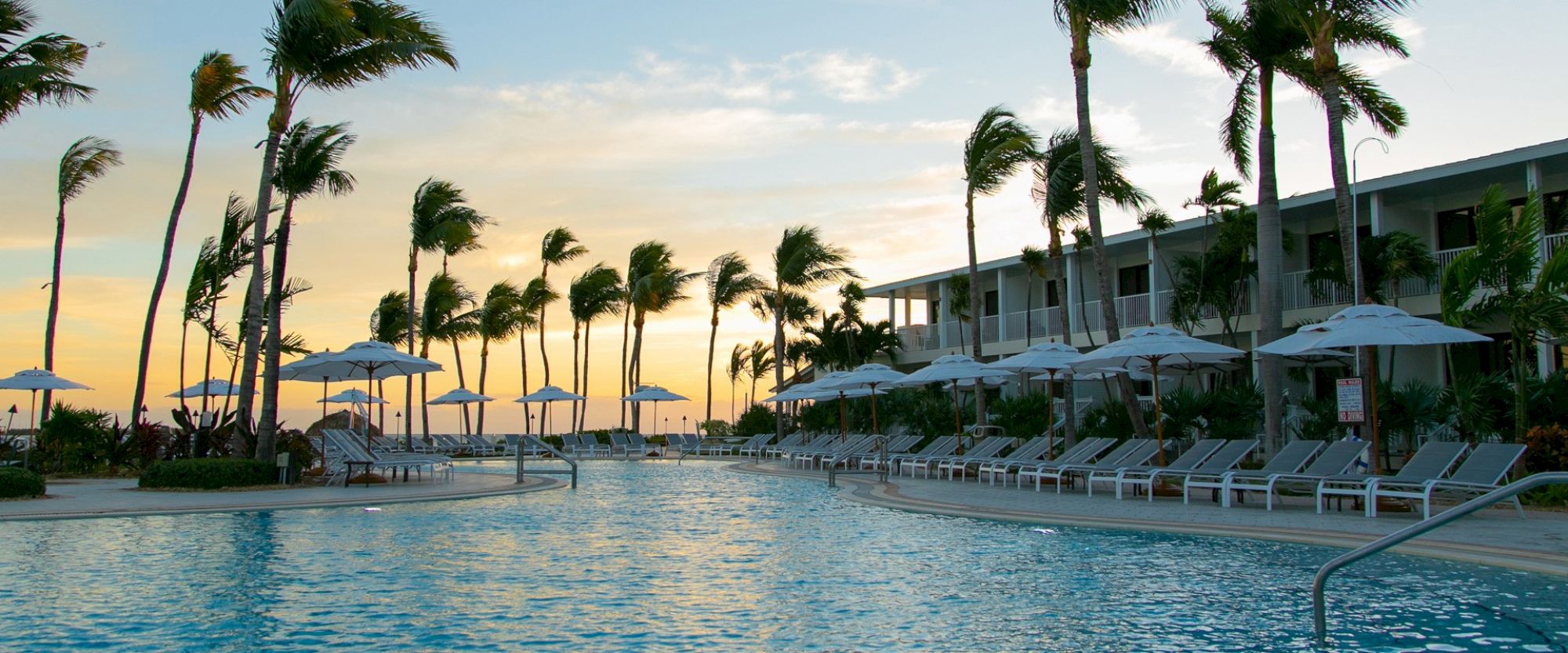 A serene resort poolside with lounge chairs, umbrellas, and palm trees at sunset, adjacent to a building.