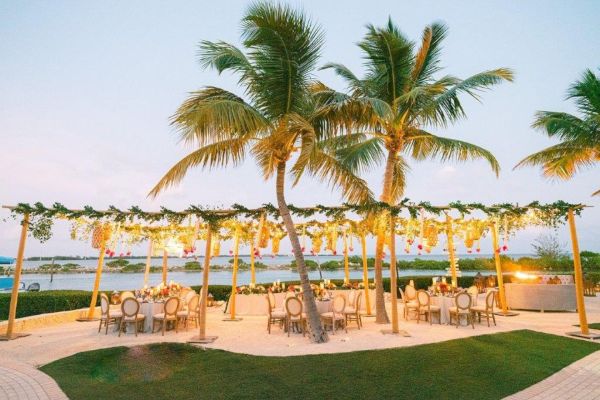 An outdoor dining setup under palm trees with string lights and decorative flowers, overlooking a body of water at sunset.