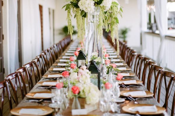 The image shows a long, elegantly set dining table with floral centerpieces, place settings, and chandeliers hanging above, likely at an outdoor event.