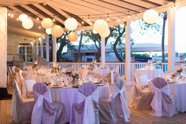 An outdoor event venue is decorated with white tables, chairs adorned with sashes, string lights, and lanterns creating an elegant ambiance at dusk.