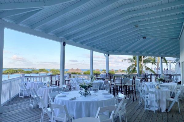 An outdoor space with white tables and chairs set up for an event, overlooking a scenic view of greenery and water under a light blue, wooden roof.